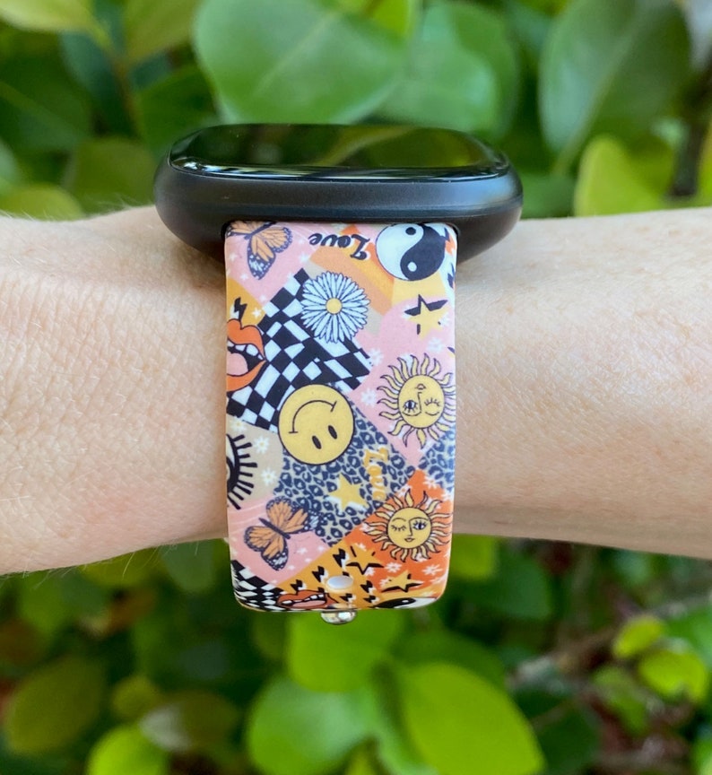 Op de afbeelding: Een roze en oranje gestreept horlogebandje met verschillende ontwerpen, waaronder een smiley, een vlinder, een zon en een yin yang symbool. Het bandje heeft de tekst "Love" erop.