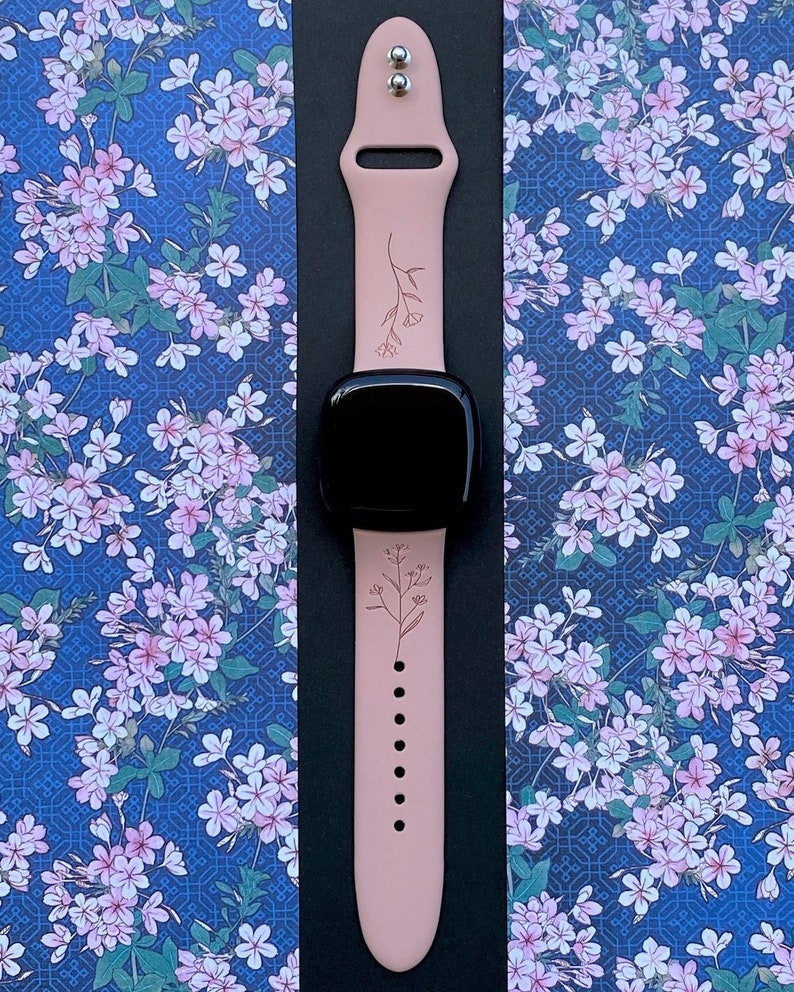 May include: A pink silicone Apple Watch band with a floral design. The band has a silver buckle and is shown on a blue background with a floral pattern.
