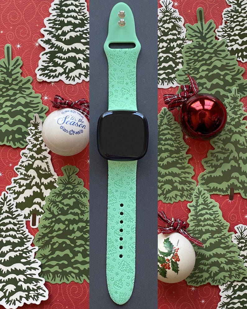 K&ouml;nnte beinhalten: Ein mintgr&uuml;nes Smartwatch-Armband mit schwarzem Bildschirm, verziert mit einem festlichen Muster aus Sternen, Weihnachtsb&auml;umen und dem Text "HO HO HO". Das Armband befindet sich auf einem roten Hintergrund mit Weihnachtbaum-Ausschnitten und Kugeln.