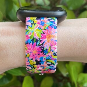 May include: A black smartwatch with a floral patterned band. The band is pink, blue, green, and yellow with a black background.