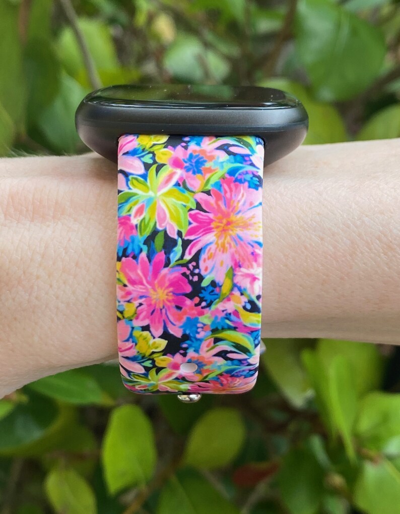 May include: A black smartwatch with a floral patterned band. The band is pink, blue, green, and yellow with a black background.