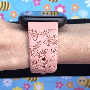 K&ouml;nnte beinhalten: Eine schwarze Smartwatch mit einem hellrosa Armband mit Blumen- und Bienenmuster. Das Armband ist mit gepr&auml;gten Blumen, Bienen und Bl&auml;ttern verziert. Die Uhr wird an einem Handgelenk vor einem Hintergrund aus Bienen und Blumen gezeigt.