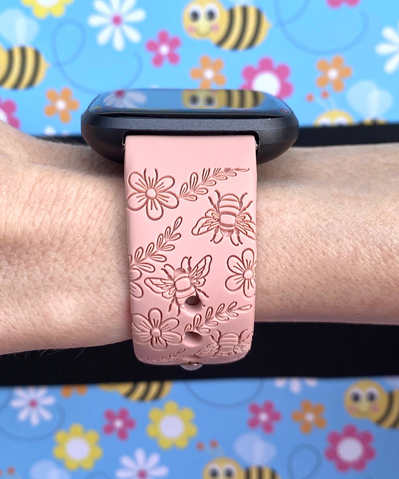 K&ouml;nnte beinhalten: Eine schwarze Smartwatch mit einem hellrosa Armband mit Blumen- und Bienenmuster. Das Armband ist mit gepr&auml;gten Blumen, Bienen und Bl&auml;ttern verziert. Die Uhr wird an einem Handgelenk vor einem Hintergrund aus Bienen und Blumen gezeigt.