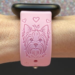 May include: Pink watch band featuring a Yorkshire Terrier dog design and three heart shapes. The band is attached to a black watch face. A small, silver-colored clasp is visible.
