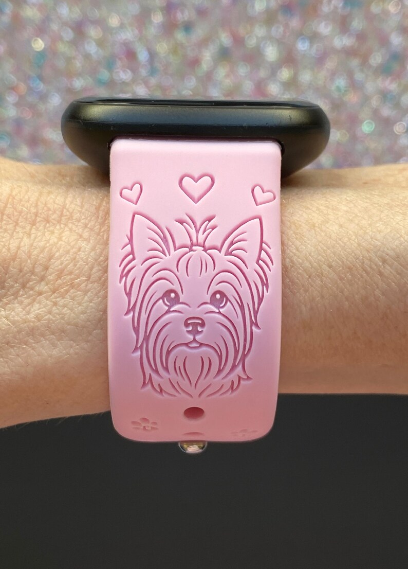 May include: Pink watch band featuring a Yorkshire Terrier dog design and three heart shapes. The band is attached to a black watch face. A small, silver-colored clasp is visible.