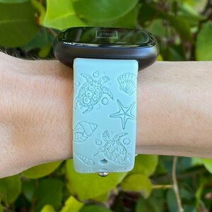 May include: A light blue watch band with a sea turtle, starfish, and seashell design. The band is made of a flexible material and is attached to a black smartwatch. The watch is being worn on a wrist.
