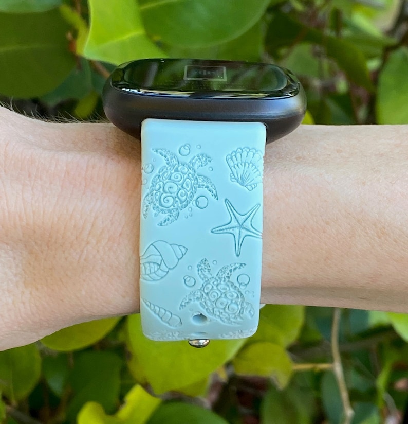 May include: A light blue watch band with a sea turtle, starfish, and seashell design. The band is made of a flexible material and is attached to a black smartwatch. The watch is being worn on a wrist.