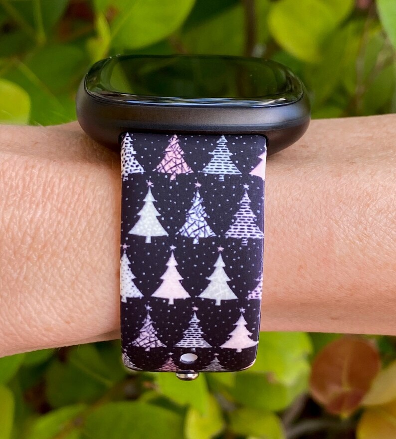 May include: A black smartwatch with a Christmas tree patterned band. The band features white, pink, and light blue trees on a black background.