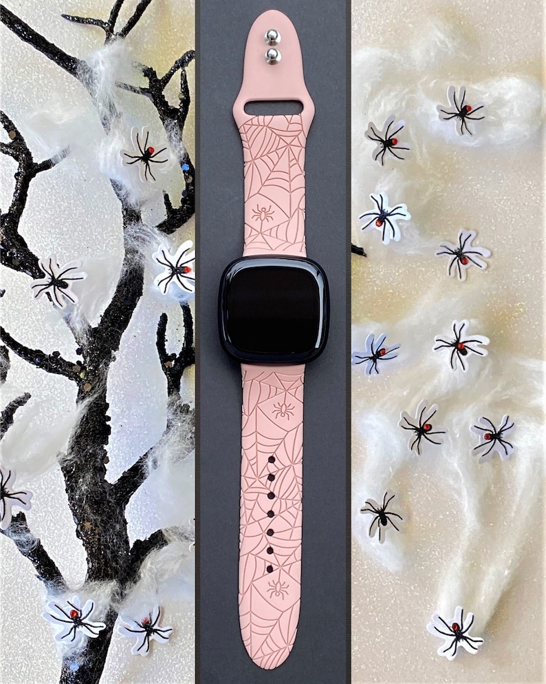 May include: A pink watch band with a spiderweb design and black spiders. The band is made of silicone and has a buckle closure.