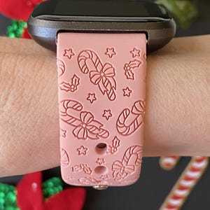 May include: A pink silicone watch band with a candy cane and holly design. The band is compatible with smartwatches.