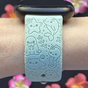 May include: A light blue watch band featuring engraved sea creatures: a starfish, dolphin, walrus, seahorse, stingray, and pufferfish. The band is adorned with floral accents and is attached to a black watch.