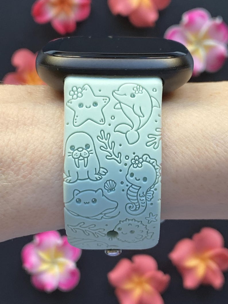 May include: A light blue watch band featuring engraved sea creatures: a starfish, dolphin, walrus, seahorse, stingray, and pufferfish. The band is adorned with floral accents and is attached to a black watch.