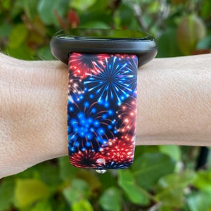 May include: Black smartwatch with a band displaying a fireworks pattern. The band has red, white, and blue bursts on a dark blue backdrop. The watch is worn on a wrist, with green foliage in the background.