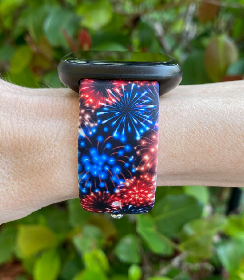 May include: Black smartwatch with a band displaying a fireworks pattern. The band has red, white, and blue bursts on a dark blue backdrop. The watch is worn on a wrist, with green foliage in the background.