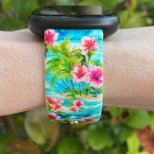 May include: A black smartwatch with a tropical floral print band. The band features pink hibiscus flowers, palm trees, and a blue ocean scene.