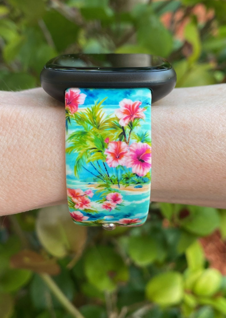May include: A black smartwatch with a tropical floral print band. The band features pink hibiscus flowers, palm trees, and a blue ocean scene.