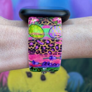 May include: A colorful watch band with a pink, green, and purple Easter egg design. The band has a leopard print pattern and lavender flowers.
