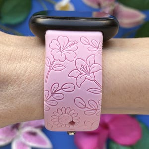 May include: A light pink smartwatch band with a floral pattern featuring hibiscus, lilies, and sunflowers. The band is attached to a black smartwatch. The design is embossed on the band. The background is a blurred blue with pink flowers.