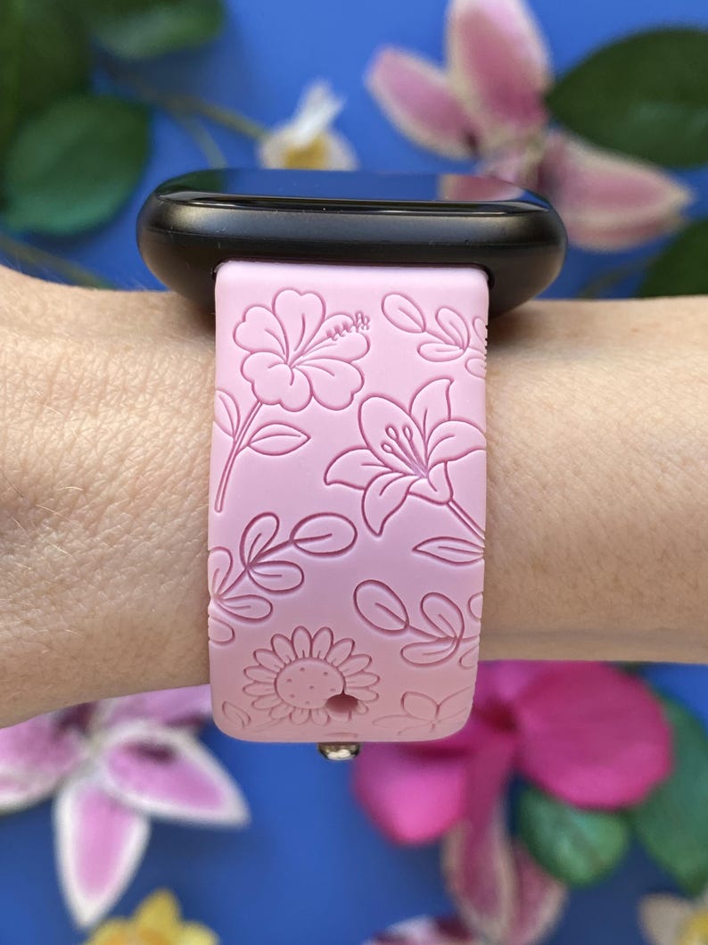 May include: A light pink smartwatch band with a floral pattern featuring hibiscus, lilies, and sunflowers. The band is attached to a black smartwatch. The design is embossed on the band. The background is a blurred blue with pink flowers.