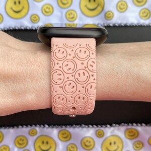 May include: A pink silicone watch band with a pattern of smiling faces. The band is on a person's wrist.