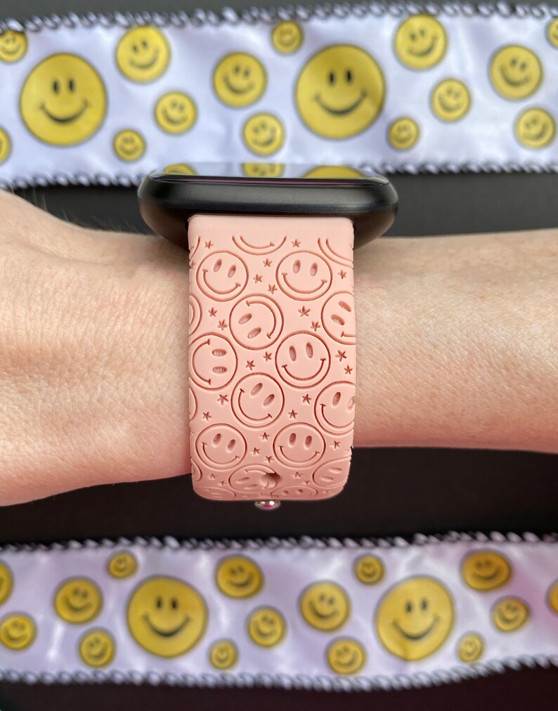 May include: A pink silicone watch band with a pattern of smiling faces. The band is on a person's wrist.
