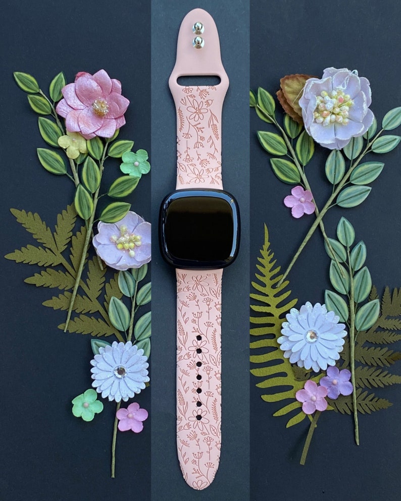 May include: A pink silicone watch band with a floral pattern. The band is designed for a smartwatch with a square face. The band is shown against a black background with paper flowers.