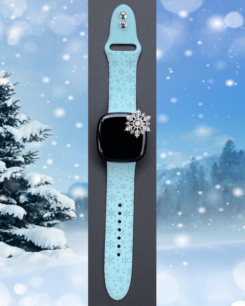 May include: A light blue silicone watch band with a snowflake pattern. The band is attached to a black smartwatch with a snowflake charm.
