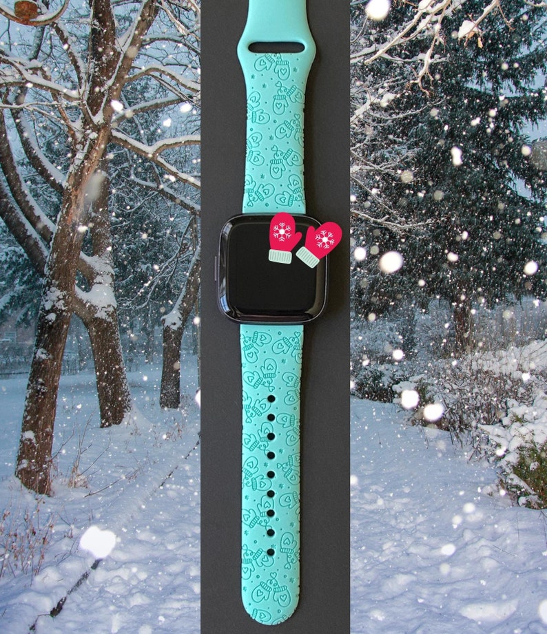 May include: A teal smartwatch band with a pattern of mittens and hearts. The watch face is black, and two red mitten-shaped charms are attached. The background shows a snowy winter scene.