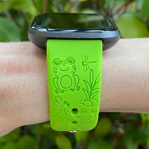 May include: A green silicone watch strap with a frog and dragonfly design. The strap is made for a smartwatch with a square face.