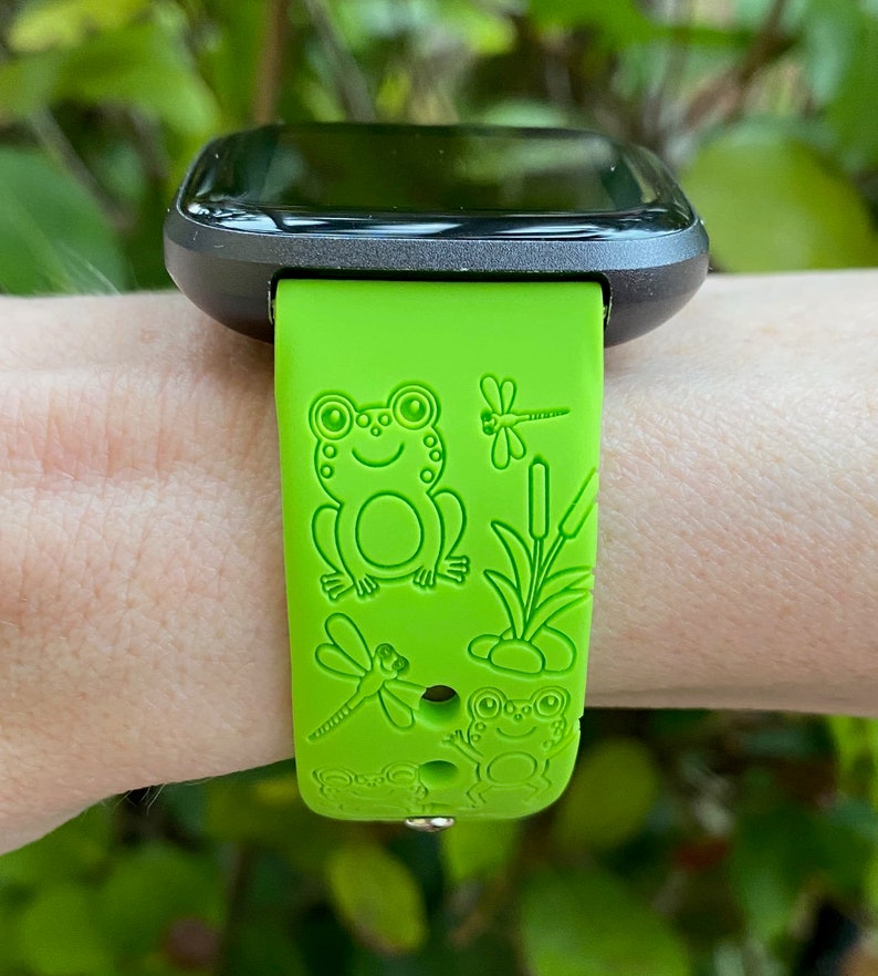 May include: A green silicone watch strap with a frog and dragonfly design. The strap is made for a smartwatch with a square face.