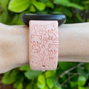 K&ouml;nnte beinhalten: Hellrosa Smartwatch-Armband mit einem sich wiederholenden Muster aus Cartoon-Katzen und Pfotenabdr&uuml;cken. Das Armband ist an einer schwarzen Smartwatch befestigt. Das Design umfasst Katzen in verschiedenen Posen und ein Fischskelett.