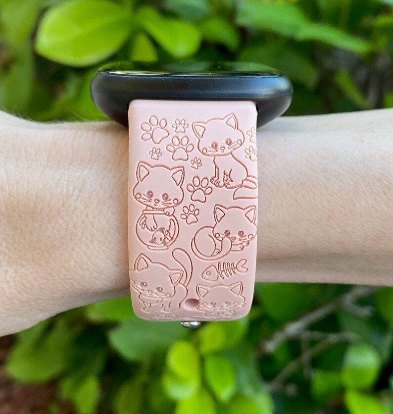 K&ouml;nnte beinhalten: Hellrosa Smartwatch-Armband mit einem sich wiederholenden Muster aus Cartoon-Katzen und Pfotenabdr&uuml;cken. Das Armband ist an einer schwarzen Smartwatch befestigt. Das Design umfasst Katzen in verschiedenen Posen und ein Fischskelett.