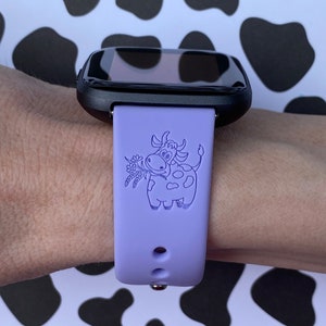 Peut inclure: Un bracelet de montre en silicone violet avec un cadran noir. Le bracelet est d&eacute;cor&eacute; d'un motif de vache de dessin anim&eacute; avec une fleur dans sa bouche.