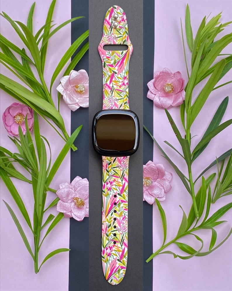 May include: A black smartwatch with a floral patterned pink and green silicone band. The band has a white background with pink, green, and yellow floral designs.
