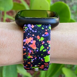 Puede incluir: Un reloj inteligente negro con una banda de dise&ntilde;o de pintura salpicada de colores. La banda tiene salpicaduras de pintura verde, naranja, rosa y p&uacute;rpura sobre un fondo negro.