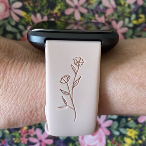 May include: A light pink smartwatch band with a floral design. The band is attached to a black smartwatch. The floral design is a simple line drawing of a flower with a stem and leaves. The background is a floral pattern.