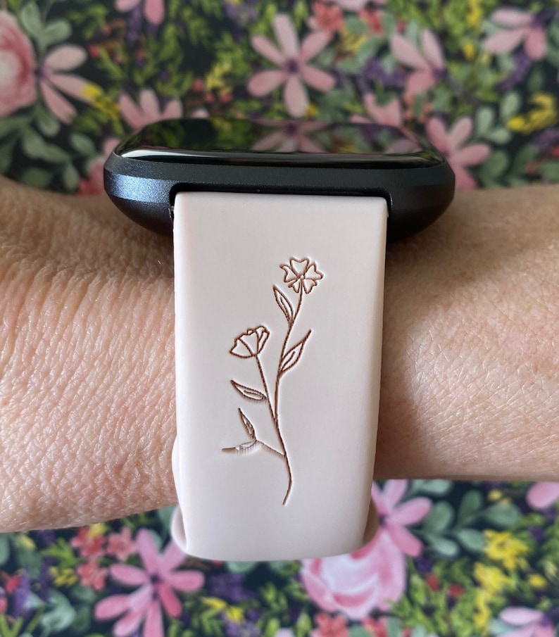 May include: A light pink smartwatch band with a floral design. The band is attached to a black smartwatch. The floral design is a simple line drawing of a flower with a stem and leaves. The background is a floral pattern.