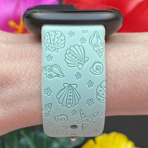 May include: A light blue silicone watch band with a seashell and starfish pattern. The band is designed for a smartwatch.