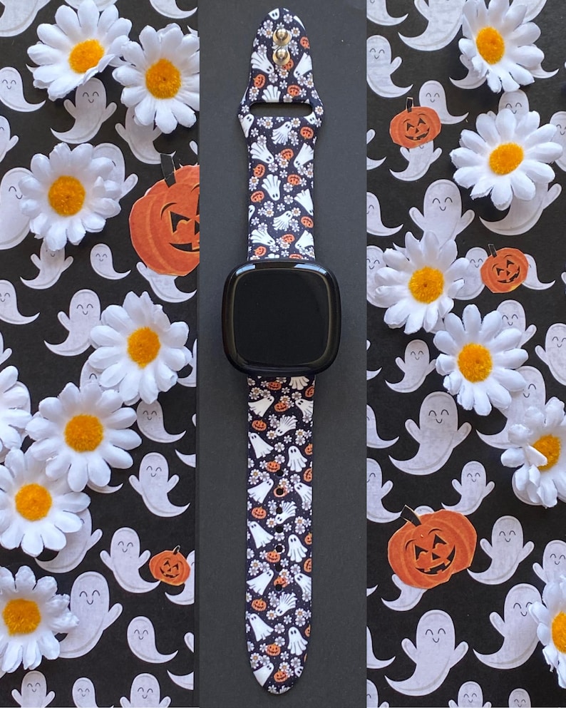 May include: Black smartwatch with a Halloween-themed band featuring white ghosts, orange pumpkins, and small flowers on a dark background. The band is made of a flexible material and is designed to fit a smartwatch. The background includes white daisies and ghost cutouts.