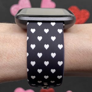 May include: A smartwatch with a black case and a black band patterned with white hearts. The watch is worn on a wrist, with pink and red heart-shaped decorations in the background.