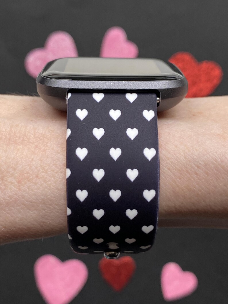 May include: A smartwatch with a black case and a black band patterned with white hearts. The watch is worn on a wrist, with pink and red heart-shaped decorations in the background.
