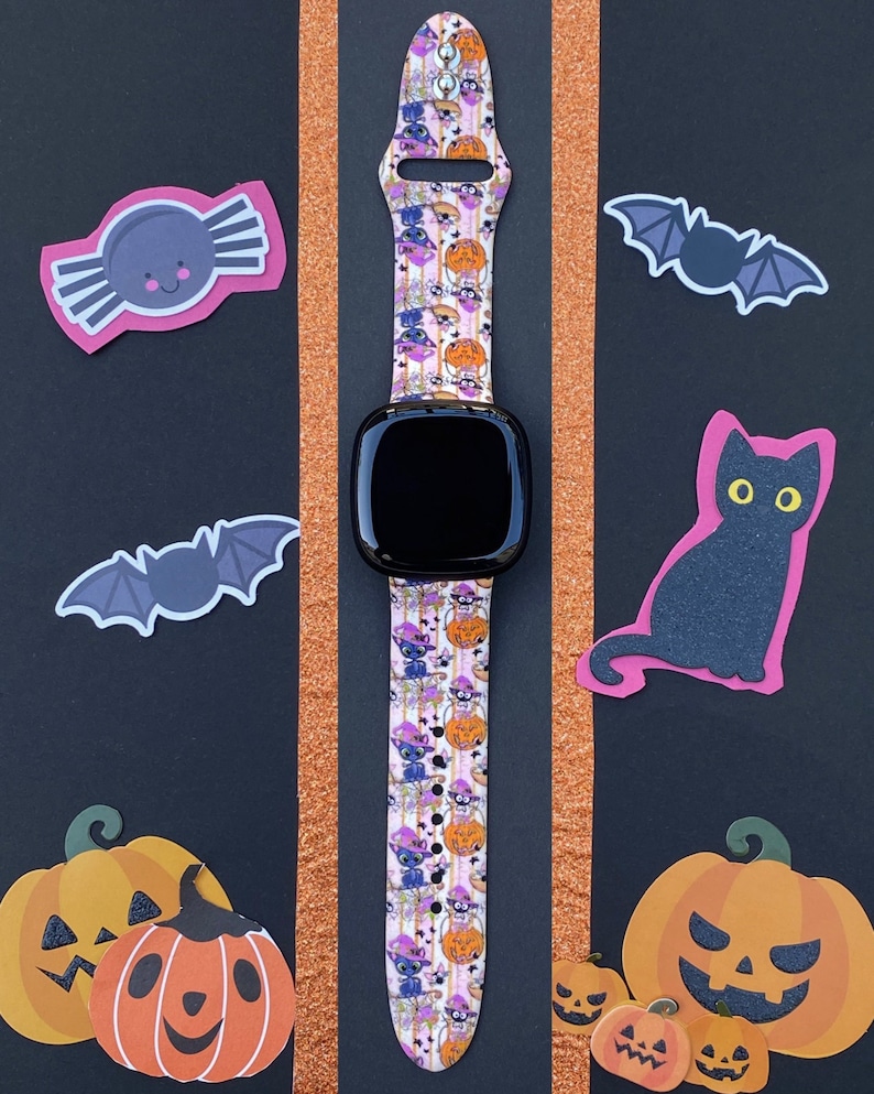 以下が含まれることがあります： ピンクとオレンジのハロウィーンをテーマにしたバンドが付いた黒いスマートウォッチ。バンドには、猫、カボチャ、コウモリ、キャンディーコーンが繰り返しパターンで描かれています。