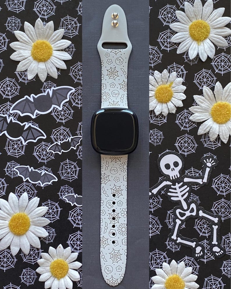 May include: A light gray smartwatch band with a black watch face, decorated with a repeating pattern of skulls, spiderwebs, and flowers. The background features daisies, bats, and a skeleton on a black background.