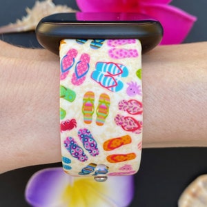 May include: A white watch band with a colorful print of flip flops, crabs, and seashells. The band is made of silicone and is compatible with Apple Watch.