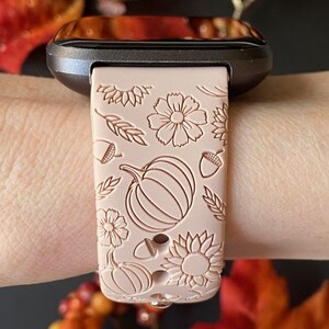 May include: A blush pink silicone watch band with a fall-themed design. The band features pumpkins, sunflowers, acorns, and wheat stalks.