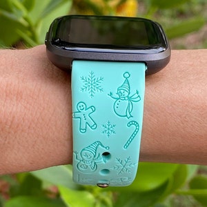 May include: A turquoise blue smartwatch band with a winter holiday theme. The band features a snowman, gingerbread man, candy cane, and snowflakes.