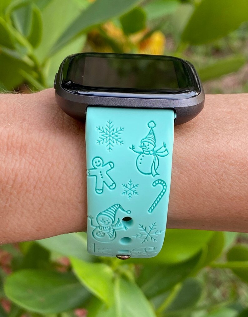 May include: A turquoise blue smartwatch band with a winter holiday theme. The band features a snowman, gingerbread man, candy cane, and snowflakes.