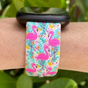 May include: A black smartwatch with a white and pink flamingo print band. The band features pink flamingos, green leaves, and yellow pineapples on a white background.