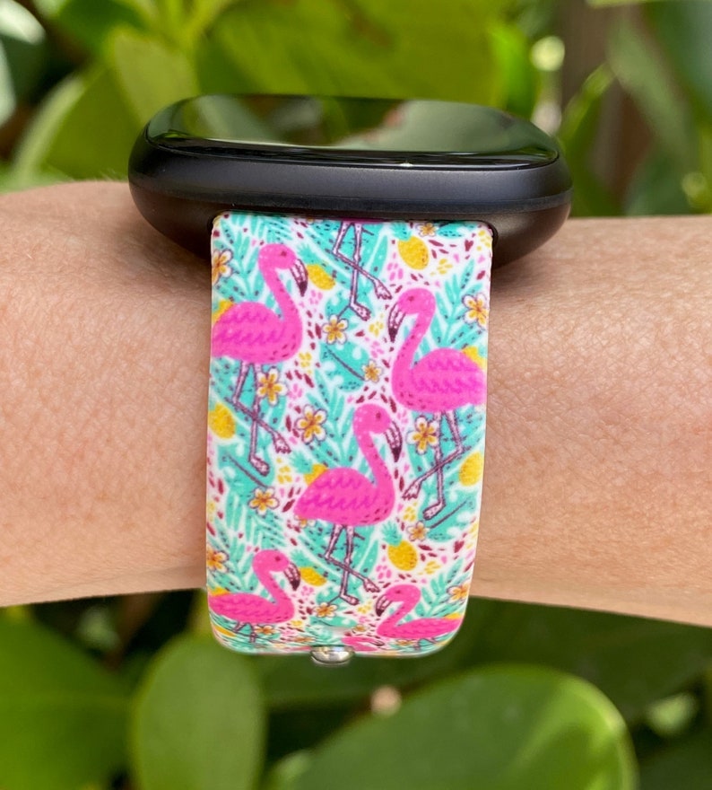 May include: A black smartwatch with a white and pink flamingo print band. The band features pink flamingos, green leaves, and yellow pineapples on a white background.