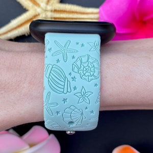 May include: A light blue silicone Apple Watch band with a seashell and starfish design. The band is on a person's wrist.